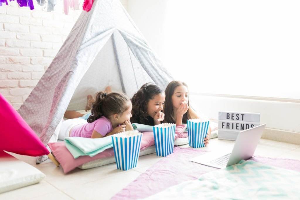 Three girls lie on the floor inside a tent, munching on popcorn and watching a laptop. A sign beside them reads "Best Friends." It's the perfect teepee setup for an unforgettable kids party rental.