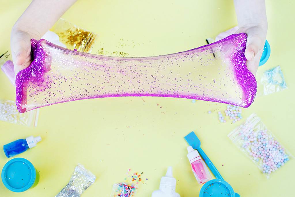 Two hands stretch a piece of pink and purple glittery slime over a table, surrounded by various craft supplies, creating the perfect scene for a kids party rental.