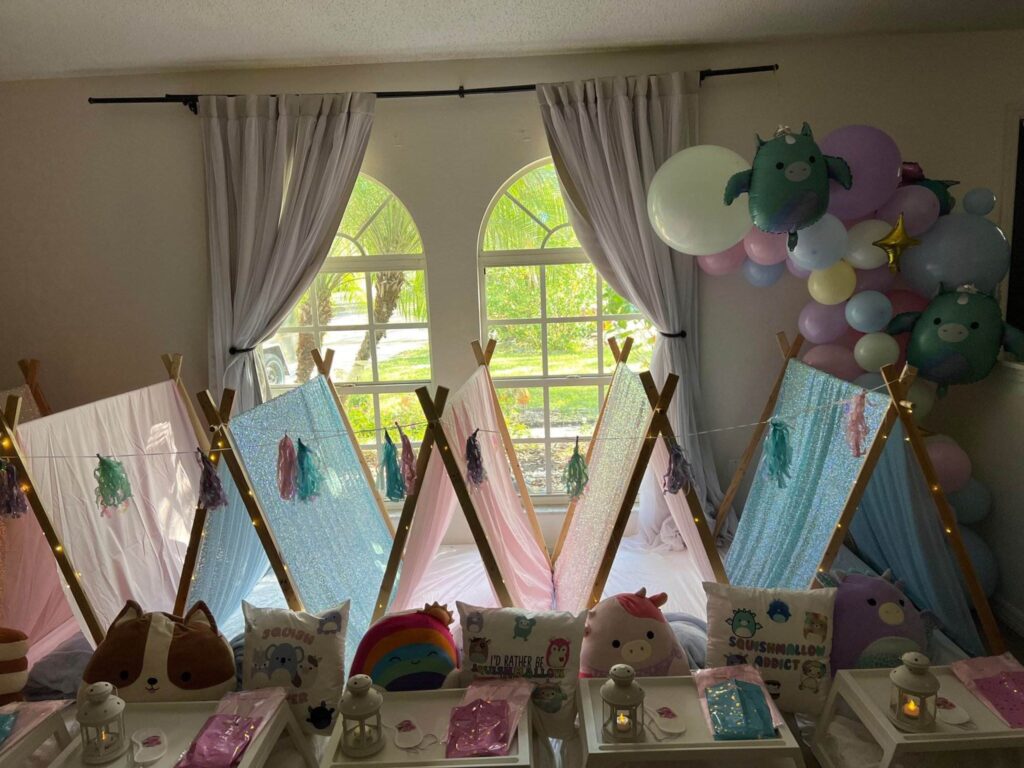 A room setup for a children's sleepover features small tents, colorful decorative pillows, and fairy lights. Perfect for a slumber party, the space is adorned with balloons and curtains framing a large window in the background. Consider this charming scene for your next kids' party rental.