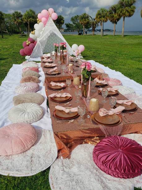 A picnic setup with a long table in a meadow. The table has pink and beige decor, glassware, plates, candles, flower vases, and is surrounded by round cushions. Perfect for a kids party rental or any event needing party service. A small canopy is in the background.