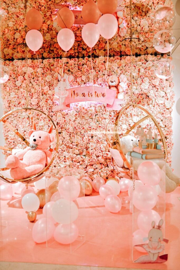 A decorative setup with a "Noah is two" banner, pink balloons, teddy bears on hanging chairs, and a floral wall backdrop could be the perfect choice for your next party rental.