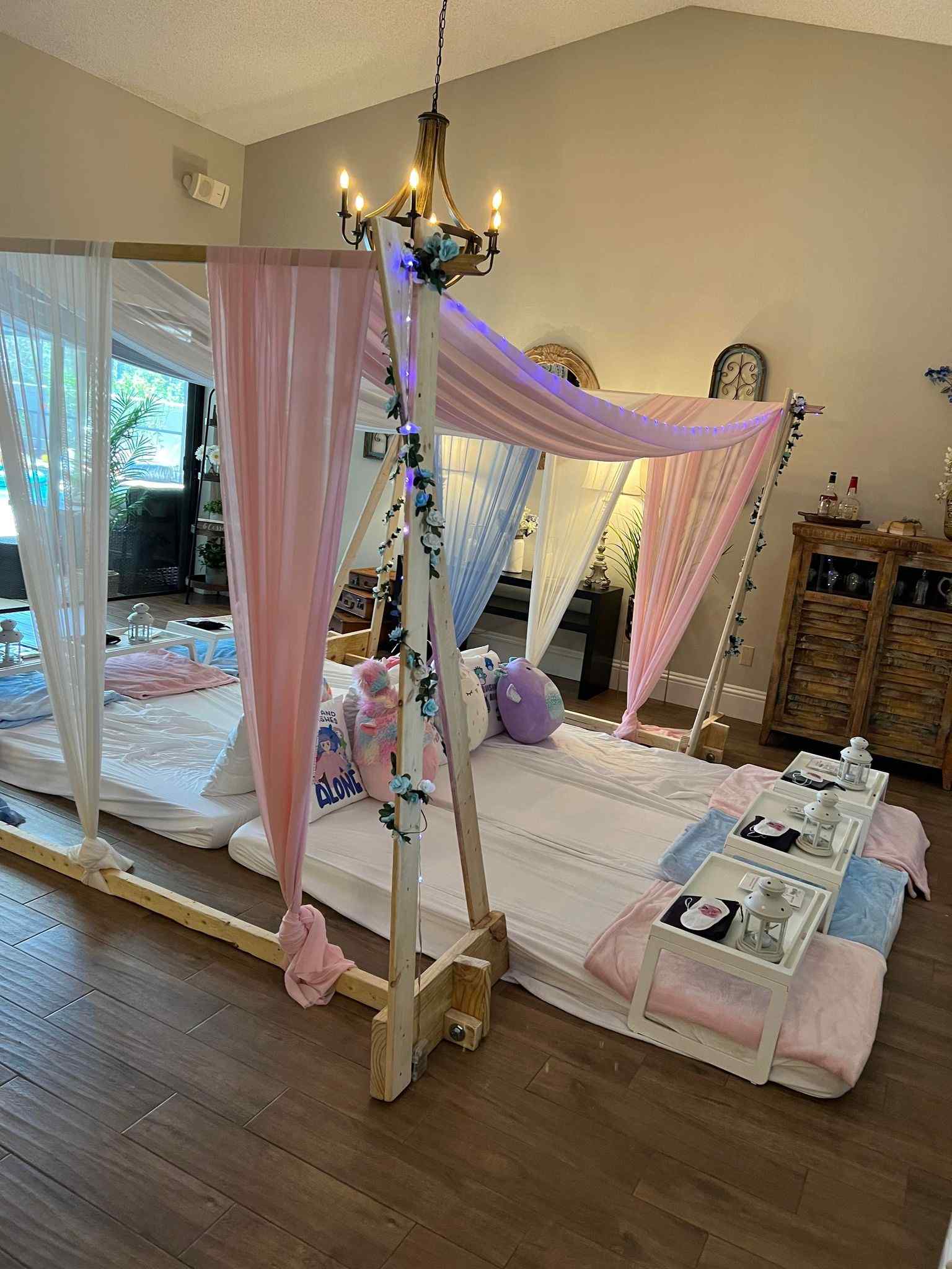 A cozy indoor canopy bed adorned with pink and white curtains, draped lights, and plush pillows, with a serving tray holding beverages beside it for the ultimate slumber party.