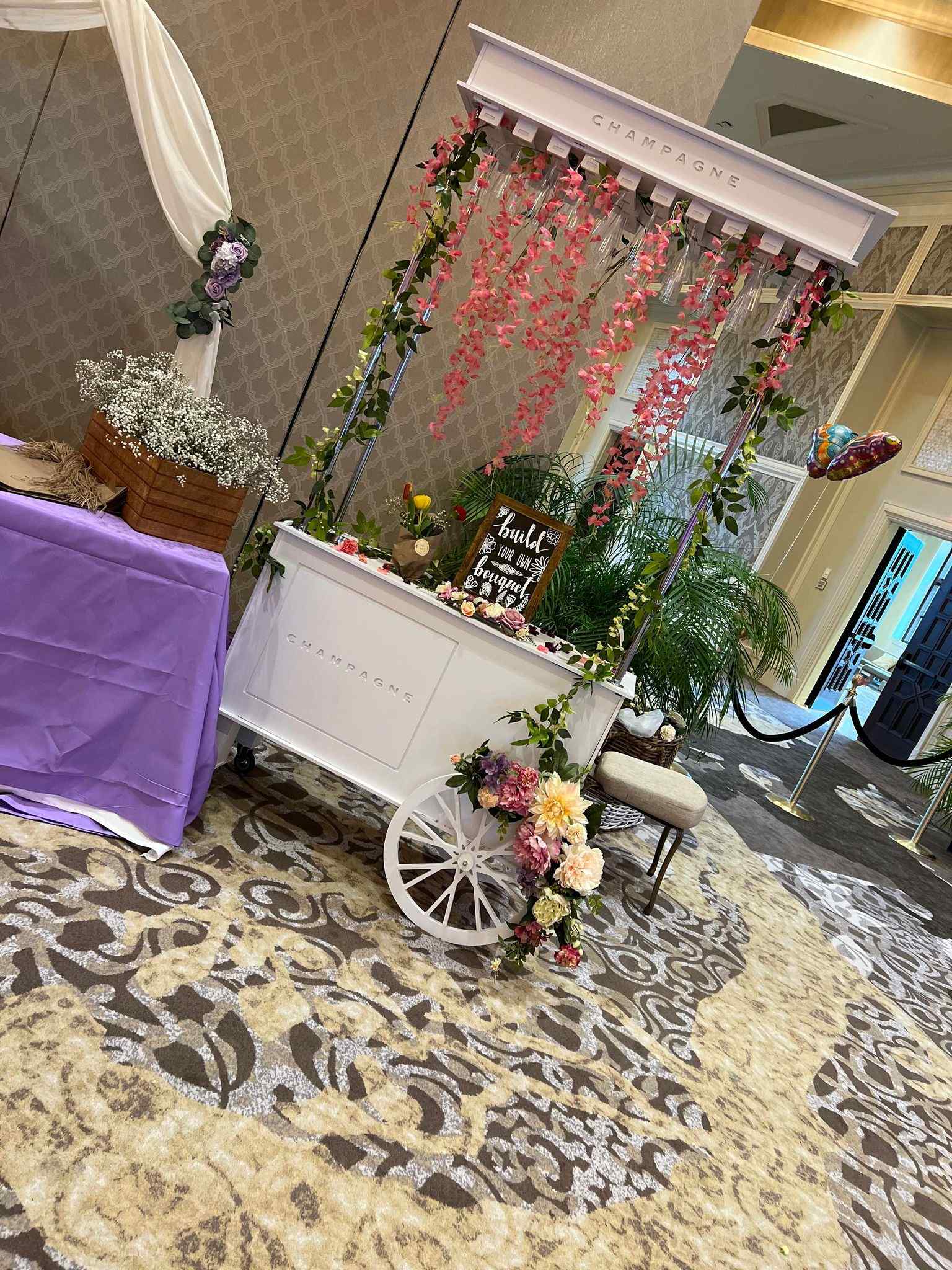 A decorated champagne cart with floral arrangements and a "champagne" sign, set up in a carpeted room with a 2024 Ultimate Sleepover backdrop.