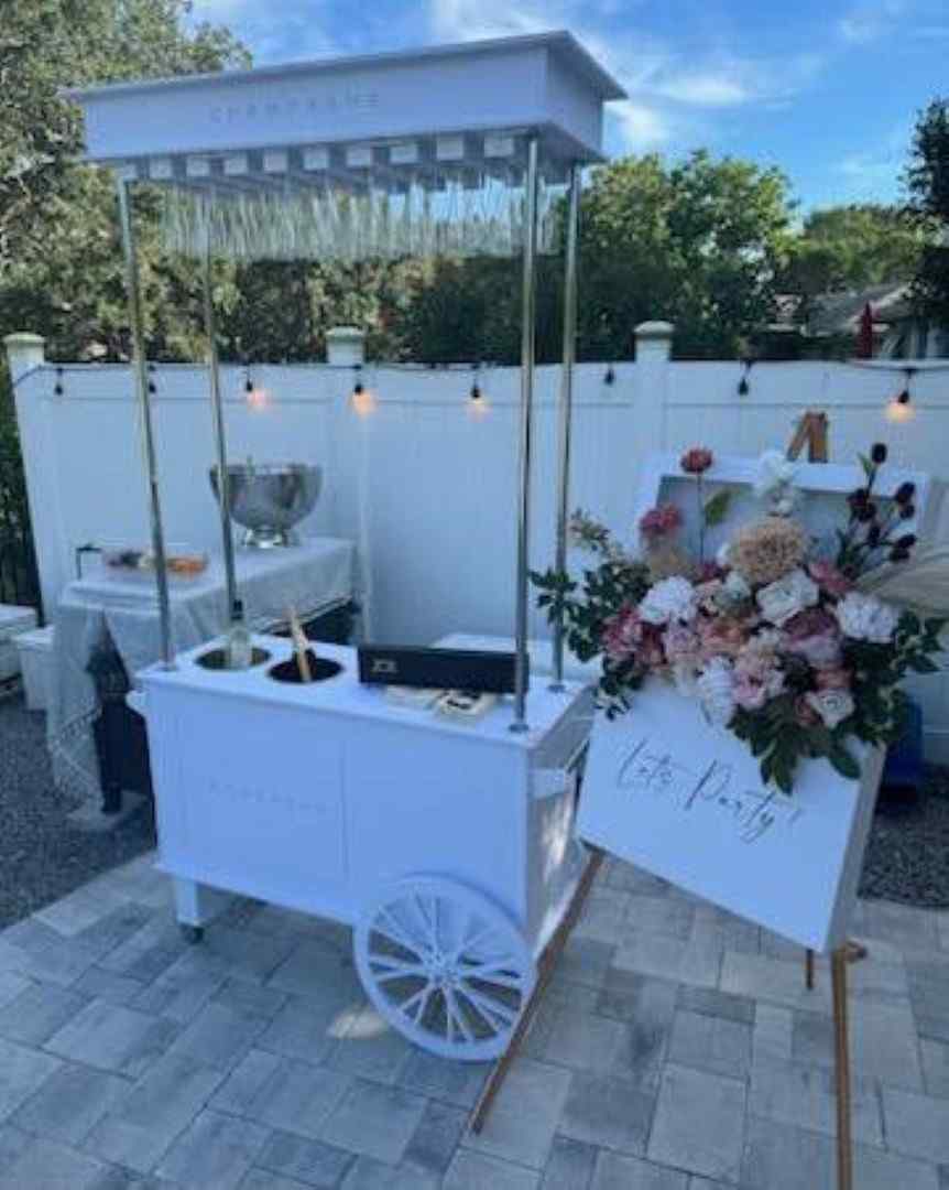 Elegant white beverage cart with a glass canopy, set up outdoors with a floral arrangement and a chalkboard sign for an adult party, under a clear blue sky.