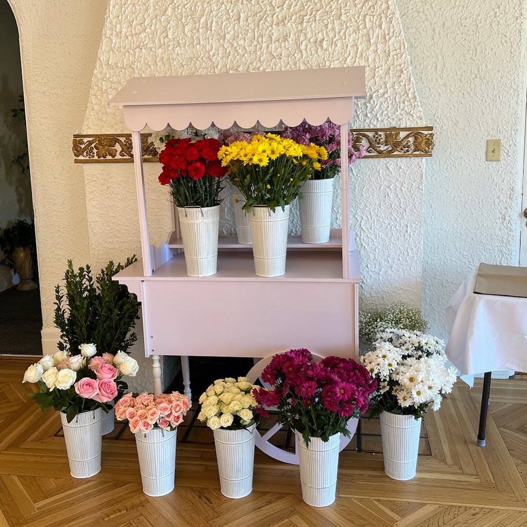 Discover the ultimate centerpiece for unforgettable baby showers: a charming pastel pink cart enhanced by a whimsical scalloped canopy. This enchanting rental option brings vibrant elegance to your event, featuring an array of colorful floral arrangements. White buckets atop the cart overflow with lush red, yellow, and purple flowers, creating a captivating display. Surrounding the cart are additional buckets brimming with delicate pink roses and pristine white daisies, beautifully arranged on a rustic wooden floor. Set against a textured white backdrop, this picturesque setup transforms any space into a floral paradise. Perfect for those seeking standout photo opportunities and dreamy event décor solutions!