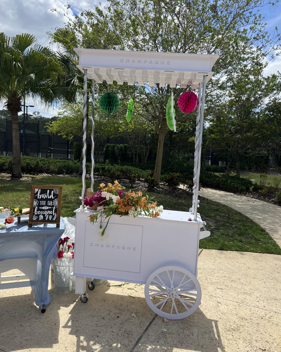 Experience the charm of a whimsical champagne cart adorned with vibrant, colorful flowers and delightful hanging decorations, all under a radiant sunny sky. Perfect for those seeking ultimate baby shower rentals, this enchanting white cart captures attention on a scenic paved path with lush trees in the background. Adding to the allure is a charming chalkboard sign inviting guests to "Build Your Own Bouquet," making it an unforgettable highlight at your outdoor celebration. Ideal for creating memorable moments and engaging with nature's beauty!