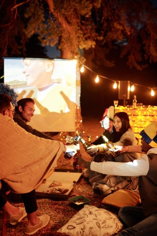 A lively group gathers under a starry night sky, illuminated by whimsical twinkling string lights as they toast with clinking bottles, savoring the enchanting outdoor atmosphere. The soothing night air complements their joyful laughter and camaraderie. Behind them, an outdoor projection screen showcases captivating scenes from Snow White, infusing the evening with a nostalgic cinematic charm and transforming the setting into a magical open-air movie experience. This perfect blend of nature, conviviality, and classic film creates an unforgettable nocturnal gathering brimming with delight and visual allure.
