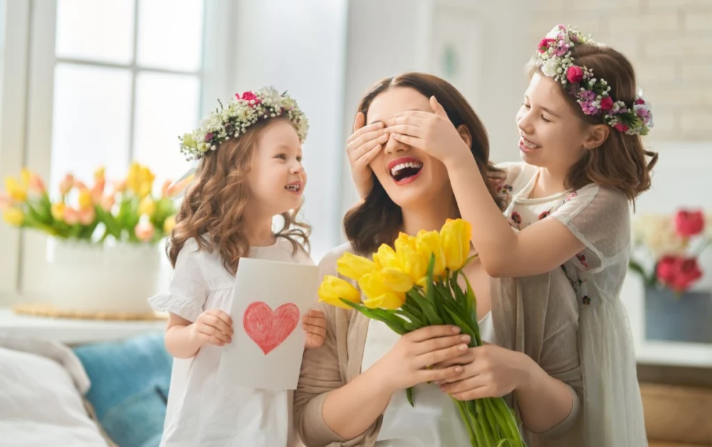 A woman holding yellow tulips is surprised by two young girls, one covering her eyes and the other holding a card with a red heart. All are smiling in light-colored clothes, ready to host a joyful Mother’s Day lounge party together.