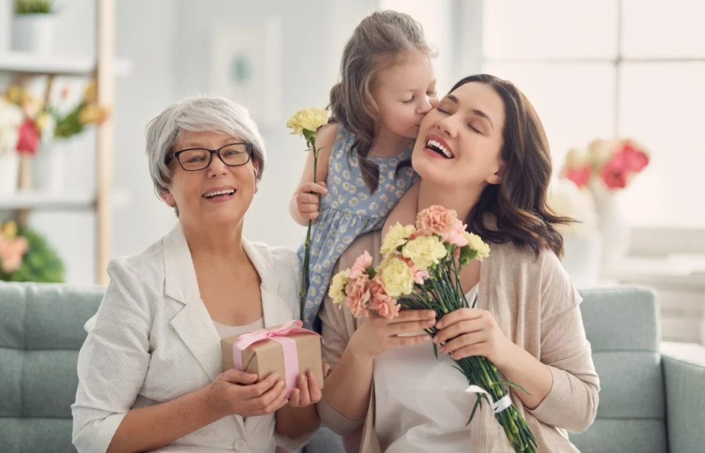 Three generations of women sit on a couch; at their Mother’s Day sleepover, a girl kisses a woman holding flowers, while an older woman holds a gift box and smiles at the camera.