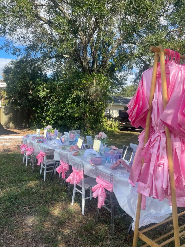 A long outdoor table is set for a party with pink decorations and gift bags—perfect inspiration for your next celebration in South Florida. Pink satin robes hang on a rack, framed by lush trees and greenery in the background, just like a trendy party shop display.