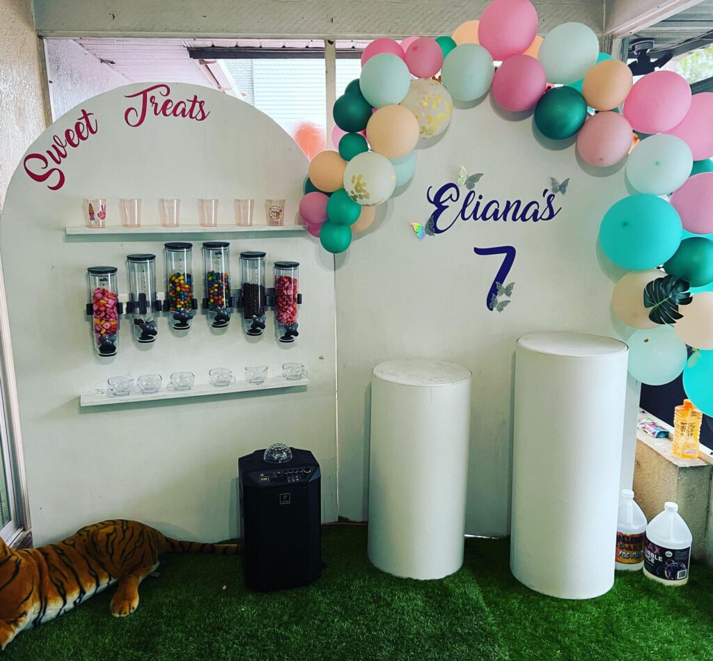 A candy display with clear dispensers, cups, and jars labeled "Sweet Treats" sits next to balloons and a sign reading "Eliana's 7" at an outdoor movie night Florida birthday party.