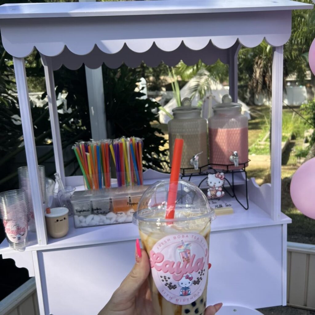 A hand holds a cup of bubble tea with a red straw in front of a decorated drink stand, offering more cups and colorful straws—perfect refreshments for an outdoor movie night in Florida.