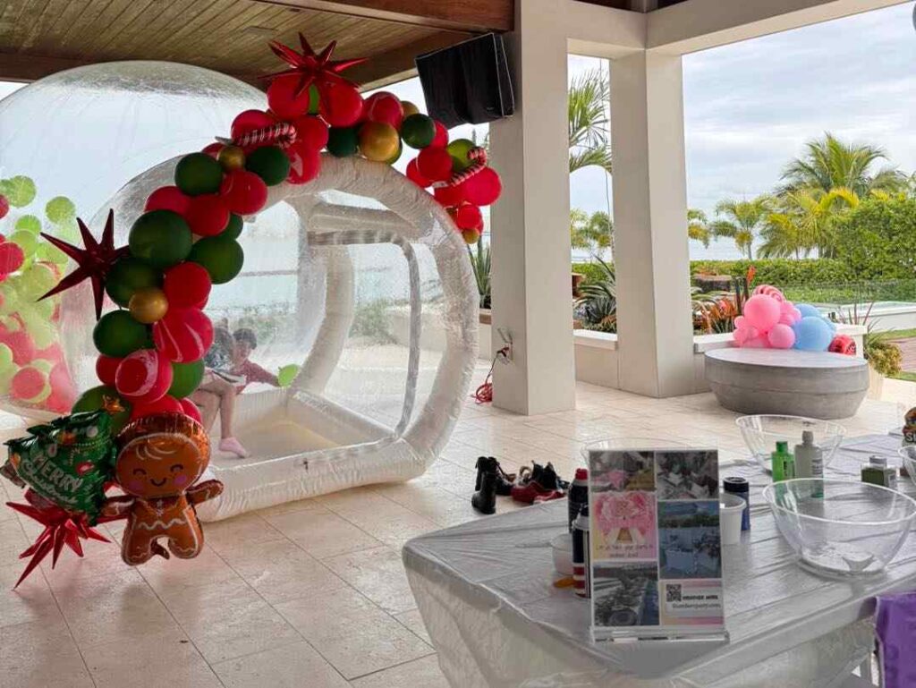 A child sits inside a transparent inflatable dome decorated with balloons, near a table with party supplies from a local party shop and a large outdoor planter with balloon sculptures in South Florida.