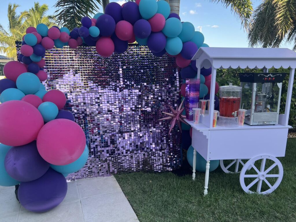 A party setup from a top party shop, featuring a sequined backdrop, colorful balloon arch in pink, purple, and teal, and a white cart with drink dispensers and cups—all beautifully arranged on the grass in Central Florida.