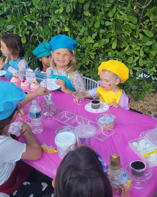 Children wearing colorful chef hats sit at a table decorated with a pink tablecloth, enjoying cupcakes and drinks at an outdoor kids activity party.
