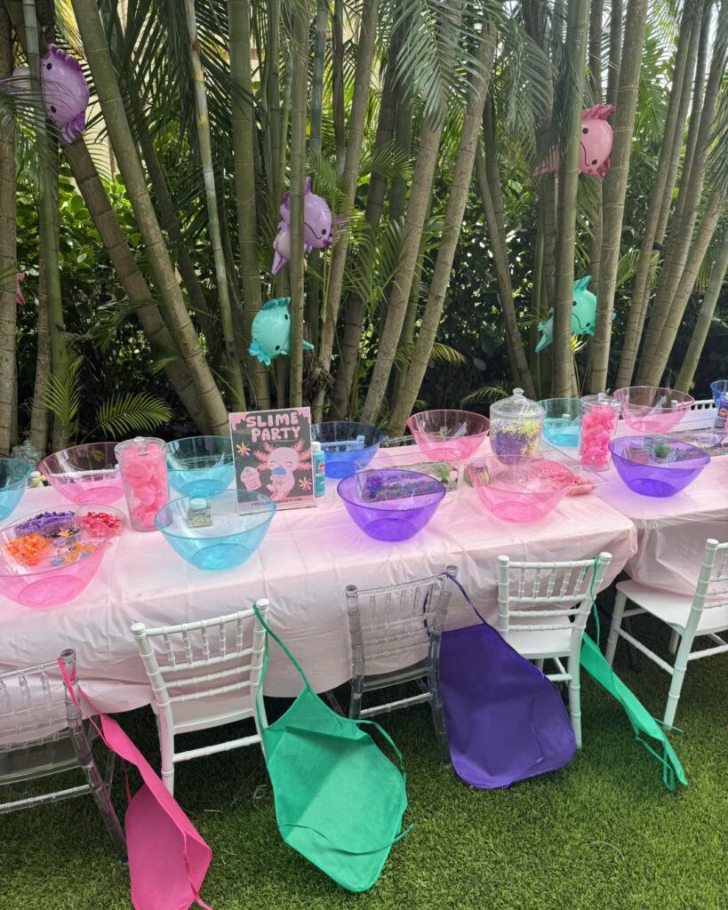 A long table outdoors is set up for a slime party by a South Florida party shop, with colorful bowls, materials, aprons on white chairs, and plush toys hanging from palm trees in the background.