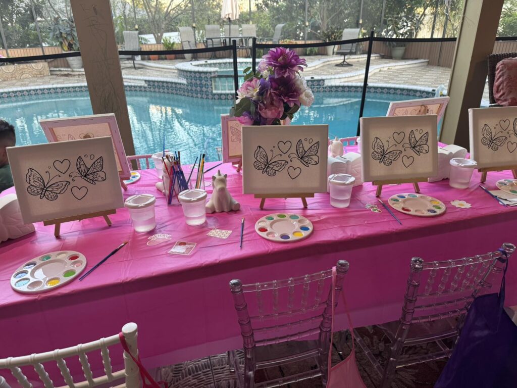 A pink-covered table by a pool is set up for a fun kids activity, featuring butterfly coloring canvases, paint palettes, brushes, and water cups ready for a creative painting session.