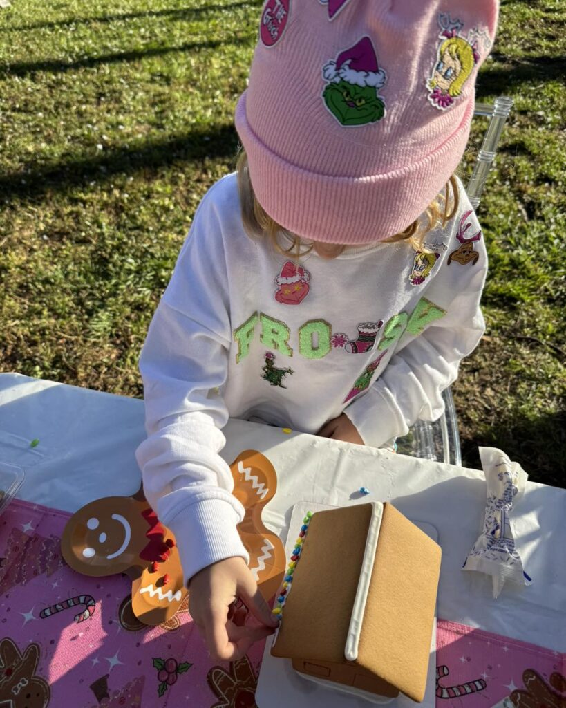 A child wearing a pink beanie and white sweatshirt enjoys a fun kids activity, decorating gingerbread cookies and a gingerbread house at an outdoor table.