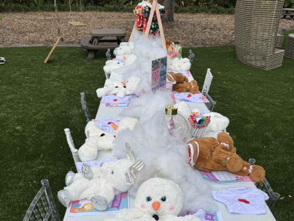 A long outdoor table is set with plush snowmen, art supplies, and white decorative fluff, evoking a festive children's craft activity—perfect for an outdoor movie night in Florida.