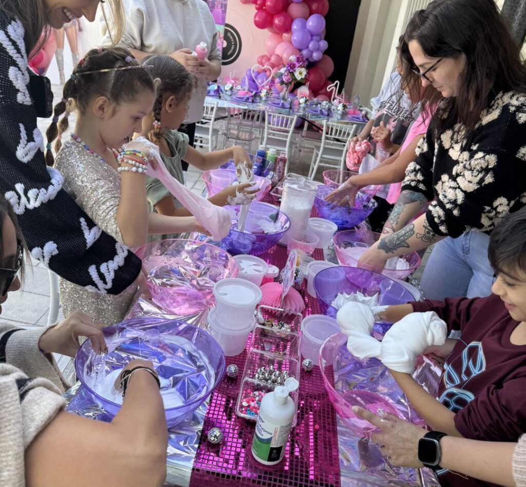 Children and adults gather around a table covered with pink decorations and bowls, enjoying a hands-on kids activitiy at a party, with cheerful balloons in the background.
