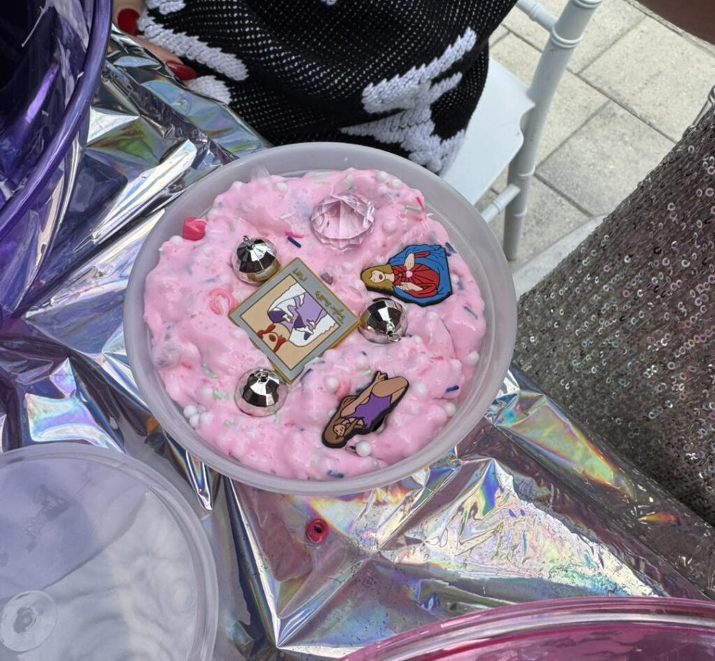 A round container filled with pink, textured slime—perfect for a kids activity—is decorated with metallic beads, a large gem, and three cartoon pins, sitting on a shiny, iridescent surface.