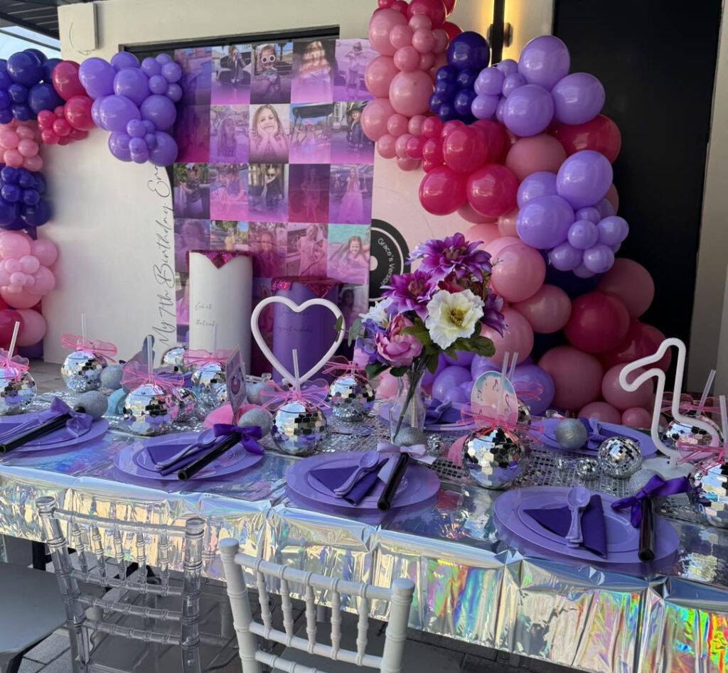 A party table with purple plates, disco ball centerpieces, and iridescent tablecloth sets a festive mood for an outdoor movie night Florida, complete with pink and purple balloons and a collage backdrop decorated with photos and flowers.