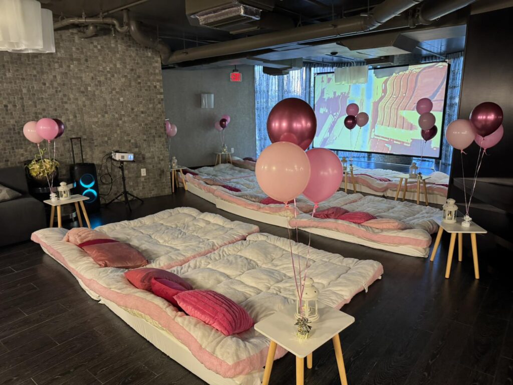 A cozy room set up like an outdoor movie night Florida style, with floor mattresses, pink and white bedding, decorative balloons, small side tables, and a projector casting images on the wall.
