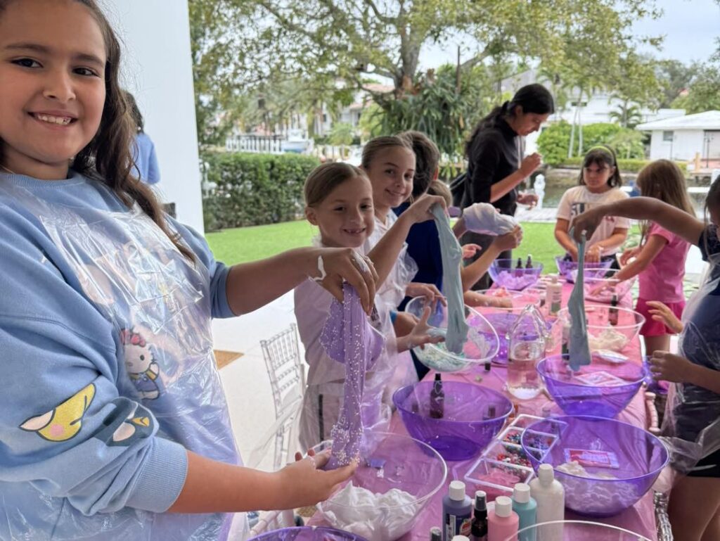 A fun kids activity unfolds as a group of children make slime at a long table filled with purple bowls, bottles, and craft supplies outdoors.
