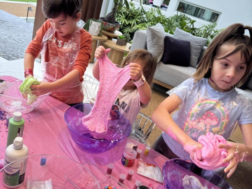 Three children enjoy a fun kids activity, making slime at a table with bowls, bottles, and craft supplies. Each child stretches or kneads a different color of slime.