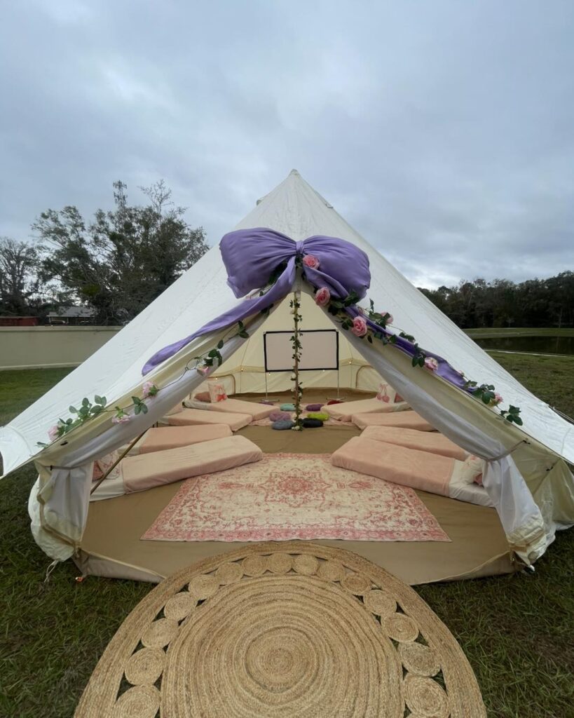 A white tent set up outdoors by a party shop in Central Florida, with a large purple bow and floral decorations at the entrance, featuring floor cushions and a rug inside on a grassy field.