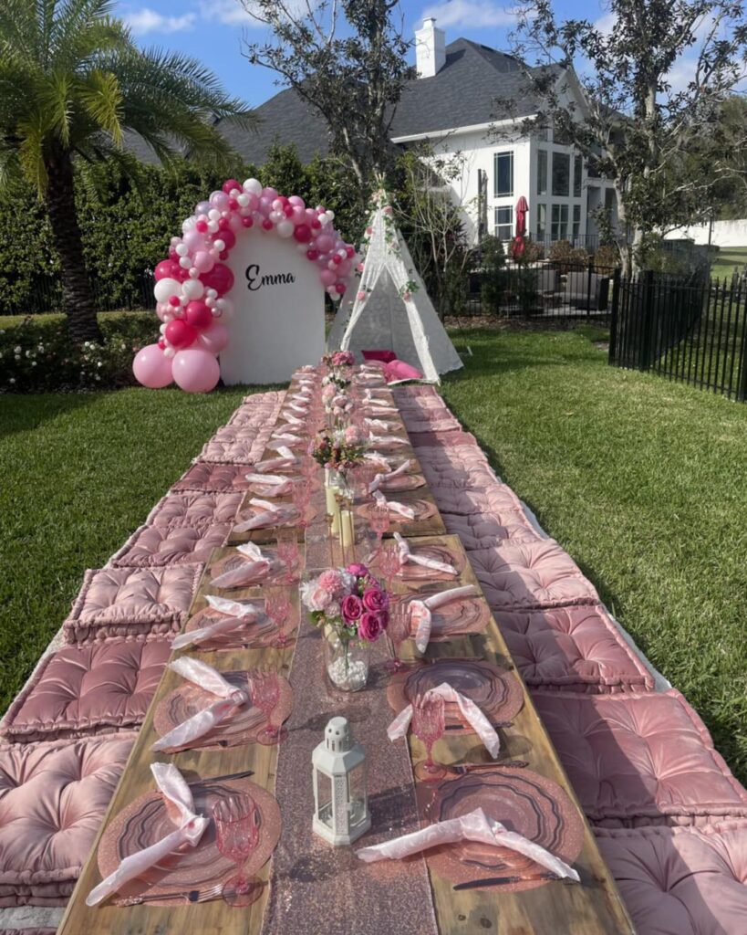 Outdoor picnic setup with a long wooden table, pink cushions, and tableware. Balloon arch and teepee tent in the background with "Emma" sign—perfect inspiration from a Central Florida party shop, set in a grassy yard.