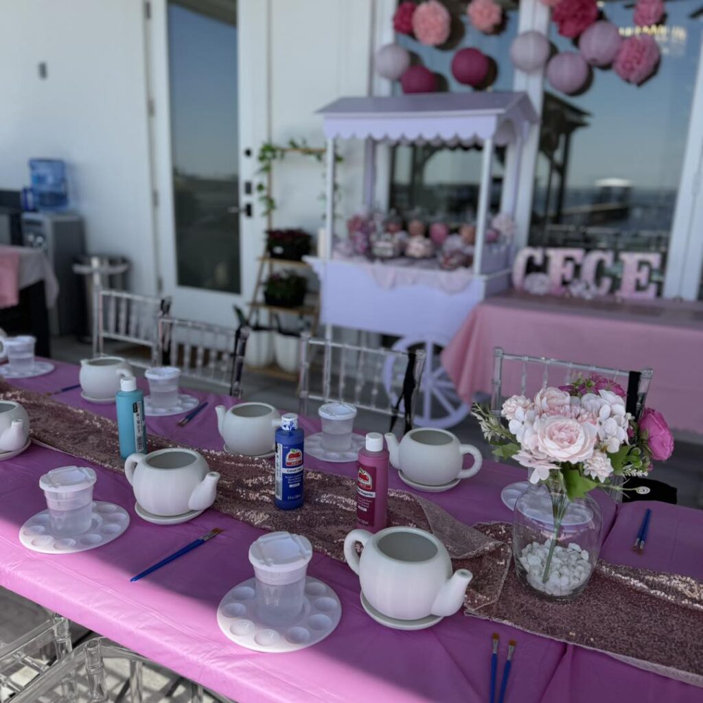 A table set for a tea party with painting supplies, teapots, cups, and a vase of flowers; a candy cart and pink decorations from a local South Florida party shop are in the background.