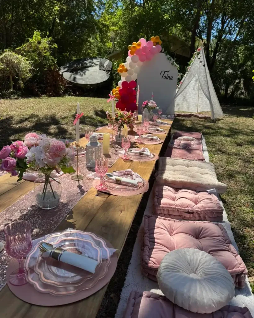 A long outdoor picnic table is set with pink and white plates, glasses, and flowers. Cushions provide seating, and a teepee with balloons stands in the background—perfect inspiration from a Central Florida party shop.