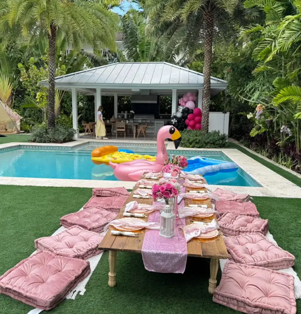 A backyard poolside setup by Slumberr, Central & South Florida's go-to party shop, features a wooden table, pink cushions, and stylish place settings; pool floats and a gazebo add flair in the background.