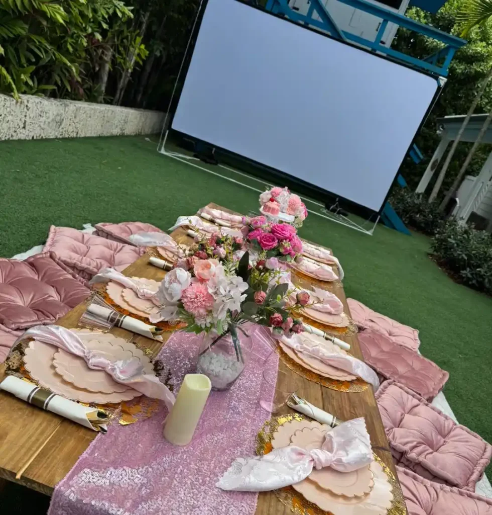 A low wooden table set for a meal with pink cushions, plates, and flower arrangements rests on the grass in front of a large movie screen—perfect for an outdoor movie night Florida style.
