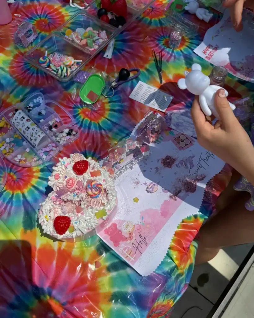 A person holds a blank bear figurine over a tie-dye table covered with art supplies, craft decorations, and a decorated dessert at a vibrant party shop in South Florida.