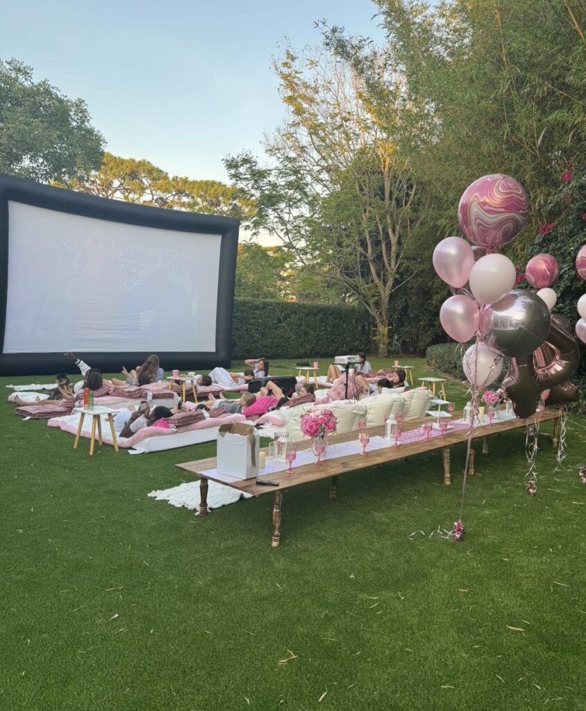 An outdoor movie night in Florida with people lounging on cushions in front of a large screen, surrounded by pink balloons and decorated tables on a grassy lawn.