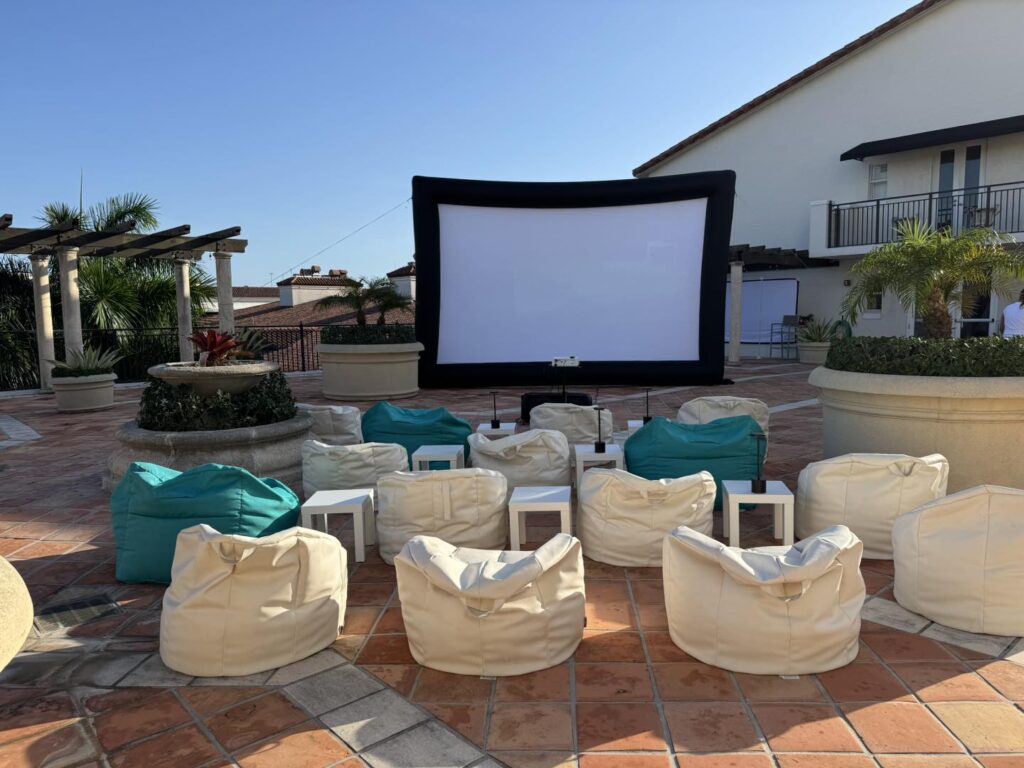 An outdoor movie night Florida setup with a large screen, white and teal bean bags, and small tables arranged on a tiled patio next to a building.