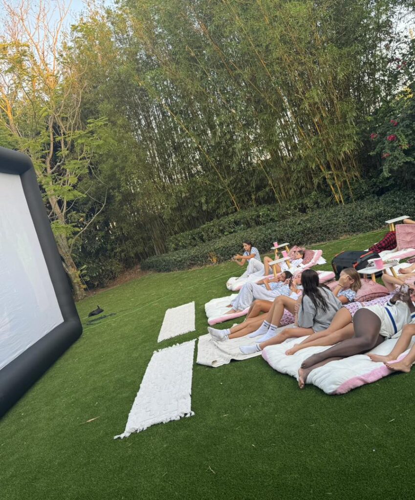 A group of people sit on blankets and pillows on grass, enjoying an outdoor movie night Florida style, watching a film on a large inflatable screen surrounded by trees and greenery.